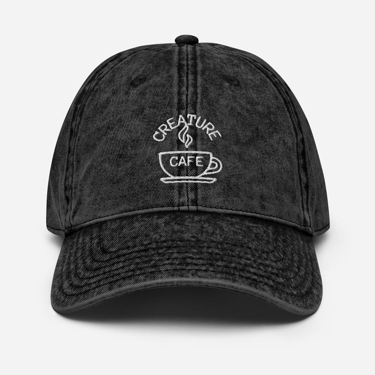 A black cotton twill cap with a logo that reads 'CREATURE CAFE' on the front, featuring a six-panel unstructured design, a black sweatband, and a metal snap buckle.